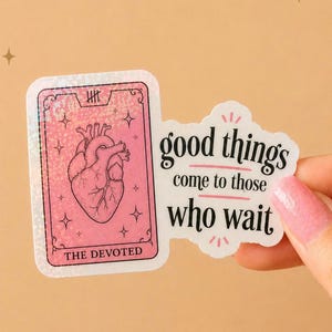 May include: A holographic sticker featuring a pink rectangular card with a heart illustration, the text THE DEVOTED, and a tally mark. Attached is a white die-cut sticker with the black text good things come to those who wait.