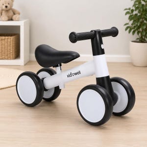 May include: A white and black balance bike with four wheels, a black seat, and black handlebars. The frame features the text "kidwell" printed in black on the side. The bike has a sturdy design with a white main body and black accents.