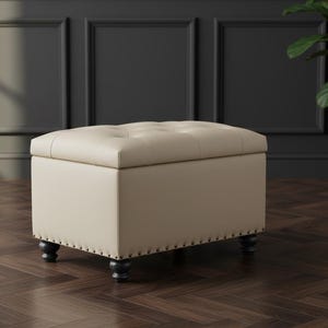 May include: A rectangular storage ottoman upholstered in a light beige faux leather material. The top surface features a button-tufted design, while the base is accented with a row of metallic nailhead trim and supported by four dark turned wooden feet.