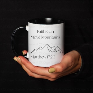 Op de afbeelding: Een witte keramische mok met een zwarte binnenkant en handvat. De mok heeft de tekst "Faith Can Move Mountains" boven een bergketenillustratie en de tekst "Matthew 17:20".