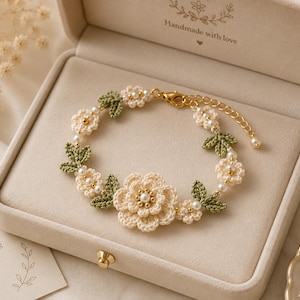 May include: A handmade crochet bracelet featuring cream-colored flowers, green leaves, and small white pearls. The bracelet is displayed in a beige jewelry box. The text "Handmade with love" is visible on the box.