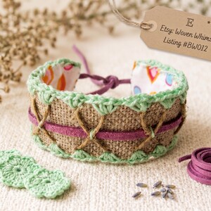 Op de afbeelding: Een handgemaakte armband met een jute basis, geaccentueerd met groene haakrand en een paarse suède band. De binnenkant heeft een kleurrijke stof met patronen. Het label luidt "Etsy: Woven Whimsy Listing #BW012".