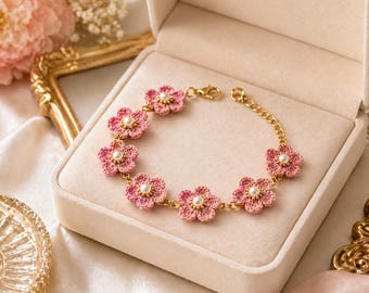 Può includere: Un delicato braccialetto a fiori all'uncinetto rosa con catena e chiusura dorate, presentato in un portagioie beige. Ogni fiore presenta un piccolo centro di perla bianca. Un accessorio affascinante.