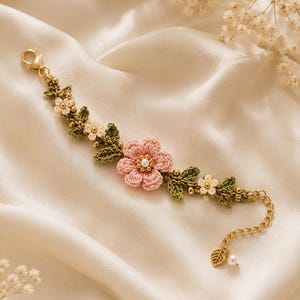 May include: A crochet bracelet featuring a central pink flower with a pearl center, flanked by green crocheted leaves and smaller white flowers. The design includes gold-toned beads, a lobster clasp, and an extender chain with a leaf charm.