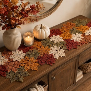 May include: A crochet table runner composed of interconnected maple leaf motifs in shades of cream, orange, red, and olive green. The runner is displayed on a wooden console table alongside a white ceramic vase, a candle, and a white pumpkin.