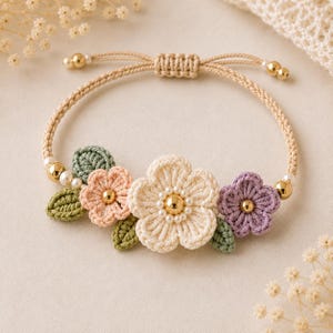 May include: A beige macrame bracelet featuring three crocheted flowers in cream, peach, and purple, with green leaves and gold beads. The bracelet has an adjustable sliding knot closure.