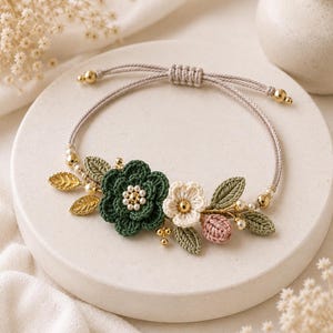 May include: A handmade bracelet featuring a floral design. The bracelet has a beige cord with gold-colored beads. The focal point is a cluster of crocheted flowers in green, white, and pink, with gold-colored leaf accents and faux pearls.