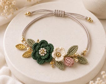 May include: A handmade bracelet featuring a floral design. The bracelet has a beige cord with gold-colored beads. The focal point is a cluster of crocheted flowers in green, white, and pink, with gold-colored leaf accents and faux pearls.
