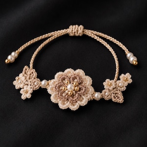 May include: A beige macrame bracelet featuring a central flower design with a pearl center and smaller floral accents. The bracelet includes small pearl and gold bead embellishments. The adjustable cord is beige.