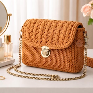 Peut inclure: Sac à bandoulière en crochet orange avec rabat et chaîne dorée. Le sac présente un motif tissé texturé et un fermoir en métal. Le fond comprend un miroir, des flacons de parfum et un vase de roses. La bandoulière mesure 100 cm.