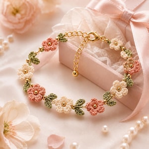 Può includere: Un delicato braccialetto floreale con fiori all'uncinetto color pesca e crema, accentuati da foglie verdi e perline dorate. Il braccialetto è ambientato su uno sfondo rosa tenue, suggerendo un tema romantico o femminile.