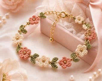 Può includere: Un delicato braccialetto floreale con fiori all'uncinetto color pesca e crema, accentuati da foglie verdi e perline dorate. Il braccialetto è ambientato su uno sfondo rosa tenue, suggerendo un tema romantico o femminile.
