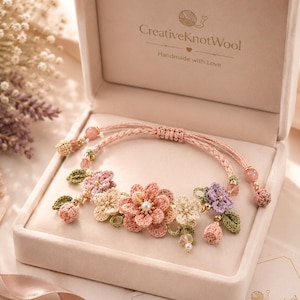 Könnte beinhalten: Ein handgefertigtes Blumenarmband in einer Geschenkbox. Das Armband zeigt gehäkelte Blumen in Rosa-, Pfirsich- und Cremetönen mit grünen Blättern und Perlenakzenten. Die Box ist hellrosa und trägt den Text "CreativeKnotWool".