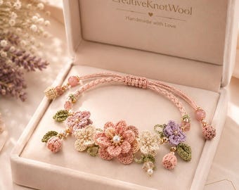 Può includere: Un braccialetto floreale fatto a mano in una confezione regalo. Il braccialetto presenta fiori all'uncinetto nei toni del rosa, pesca e crema, con foglie verdi e perle. La scatola è rosa chiaro e ha il testo "CreativeKnotWool".