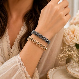 Könnte beinhalten: Zwei Armbänder mit Perlen, eines goldfarben und eines dunkelgrau, werden am Handgelenk einer Person getragen. Das goldfarbene Armband hat eine Reihe von verbundenen runden Perlen. Das dunkelgraue Armband hat kleinere, dunklere Perlen. Die Armbänder werden vor einem weichen, neutralen Hintergrund präsentiert.