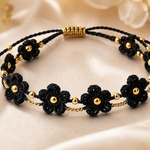 Könnte beinhalten: Ein schwarzes Blumenarmband mit goldenen Akzenten. Das Armband zeigt mehrere schwarze gehäkelte Blumen mit goldenen Zentren und goldenen Perlen. Die verstellbare Kordel ist schwarz mit goldenen Details.