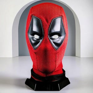 Peut inclure: Un masque intégral rouge avec une surface en tissu texturé et de grands empiècements oculaires noirs angulaires. Les zones des yeux contiennent des lentilles blanches en maille. Le masque est présenté sur un support noir.