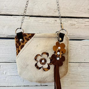 Op de afbeelding: Een kleine, crèmekleurige crossbodytas met een kettingriem. De tas heeft een luipaardprint accent op een hoek en een bloemenontwerp. Extra bloemversieringen en een kwastje hangen aan de zijkant. De tas is gemaakt van een zacht, getextureerd materiaal.