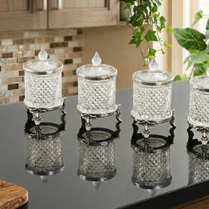 Op de afbeelding: Vier heldere glazen potten met ruitvormige zijkanten en zilverkleurige randen en deksels. Elke pot staat op een decoratieve zilverkleurige standaard met drie poten. De potten staan op een zwart aanrecht.