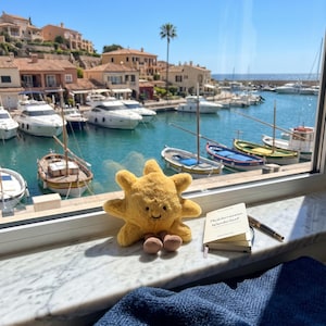 Peut inclure: Une peluche soleil jaune avec un visage souriant et de petits pieds marron, placée à côté d'un petit carnet crème portant l'inscription "Mediterranean Wanderlust" sur la couverture et d'un stylo noir, sur un rebord de fenêtre en marbre blanc.