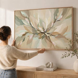 Puede incluir: Una gran impresión artística en lienzo enmarcada con un diseño floral abstracto en tonos apagados de beige, verde y marrón. La obra de arte se exhibe en una pared blanca. El marco es de madera clara.