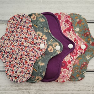 May include: Six reusable cloth menstrual pads with floral patterns in pink, purple, and green. Each pad is secured with a white snap. The pads are displayed on a white wooden surface, showcasing a variety of designs.
