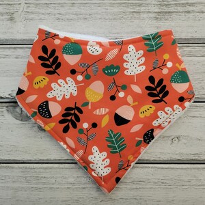 May include: A red bandana with a white, black, green, and yellow pattern of leaves and acorns. The bandana is made of soft fabric and has a white fleece lining.
