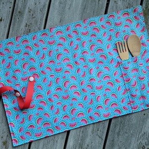 May include: A blue placemat with a watermelon pattern, featuring red and pink watermelon slices on a blue background. A pocket holds a wooden fork and spoon. A red strap with a snap closure is attached.
