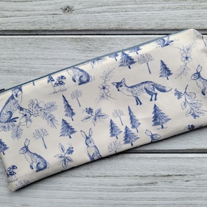May include: A white zippered pouch with a blue and white woodland pattern. The pattern features foxes, rabbits, trees, and mushrooms.