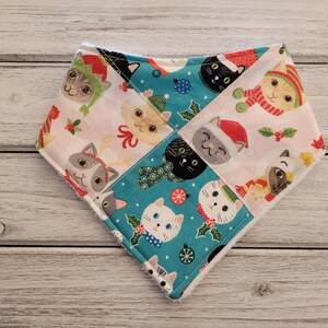 May include: A white, blue, and pink bandana with a Christmas cat pattern. The bandana is made of fabric and has a triangular shape.