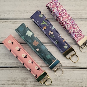May include: Four fabric keychains with different floral patterns. The keychains are pink, blue, green, and pink with white flowers. Each keychain has a gold metal ring.