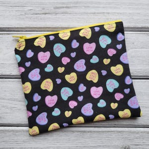 Peut inclure: Une pochette noire à fermeture éclair avec un motif répété de cœurs de bonbons colorés. Les cœurs ont différents messages, notamment "I Love You", "I Love Mew" et "Not Perfect, Just Purrfect".