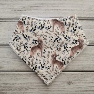 May include: A white baby bib with a repeating pattern of brown wolves and blue and gray leaves. The bib is made of soft fabric and has a rounded shape.