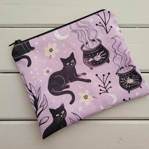 May include: A rectangular zipped pouch in light purple featuring a repeating pattern of black cats, steaming cauldrons with sun symbols, white crescent moons, small flowers, and thin leafy branches. The pouch has a black zip closure.