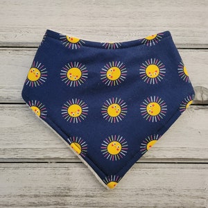 May include: A navy blue baby bib with a repeating pattern of smiling yellow suns with colorful rays. The bib is triangular and has a white, absorbent backing. A cheerful accessory for mealtime.