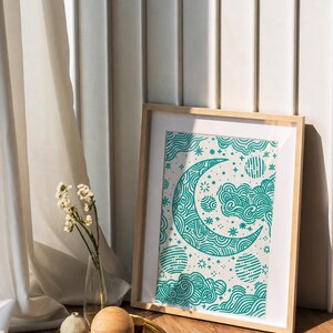 Puede incluir: Una impresión artística enmarcada con un diseño de luna creciente turquesa, estrellas y nubes sobre un fondo crema. La obra de arte está en un marco de madera clara, apoyada contra una pared blanca. Una bandeja dorada con esferas decorativas y flores está al frente.