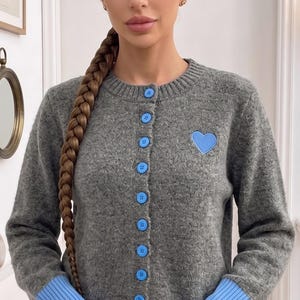 May include: A grey cardigan with a blue heart patch and blue cuffs. The button-down sweater has blue buttons and is made of a soft, textured material. The cardigan is a casual, everyday garment.