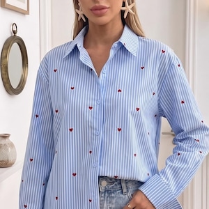 Peut inclure: Chemise à rayures bleues et blanches avec de petits cœurs rouges. La chemise à manches longues a un col et des poignets classiques. Elle est portée avec un jean bleu.