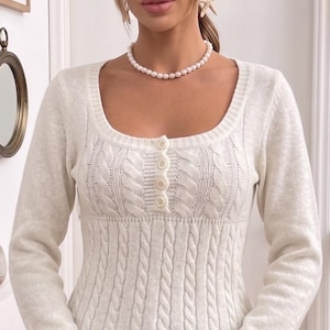Könnte beinhalten: Ein weißer Zopfstrickpullover mit rundem Ausschnitt und langen Ärmeln. Der Pullover hat eine Knopfleiste auf der Brust und eine taillierte Silhouette. Eine Perlenkette und Seestern-Ohrringe ergänzen den Look.