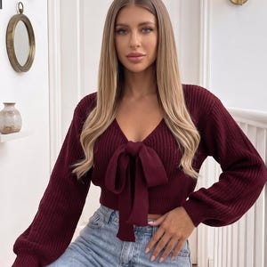 Peut inclure: Un pull en tricot bordeaux à manches longues avec une encolure en V et un nœud en tissu noué sur le devant. Le vêtement est fabriqué dans une maille côtelée avec des poignets resserrés et une coupe courte, porté avec un jean en denim bleu clair.