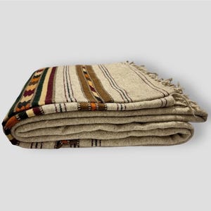 May include: A folded woven blanket in a light beige color featuring a series of vertical stripes in dark green, burgundy, brown, and orange. The fabric has a textured appearance and includes decorative fringe along the edge of the textile.