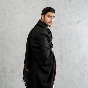 May include: A solid black shawl featuring a thin maroon border along the edge and a decorative band with gold and black geometric patterns near the fringed end. The fabric drapes over the shoulders, showcasing a smooth, matte texture.