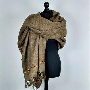 Può includere: Uno scialle marrone tessuto con un bordo a frange e dettagli ricamati decorativi in arancione, verde e giallo. Lo scialle è drappeggiato su un manichino nero, mostrando la sua consistenza e il suo design. Un accessorio caldo.