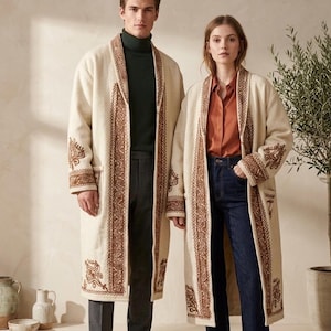 Pode incluir: Dois casacos longos abertos de cor creme com bordados castanhos intrincados nas lapelas, punhos e bolsos. Os casacos são usados sobre calças e camisas escuras, exibindo um design estruturado de comprimento total com bolsos laterais.