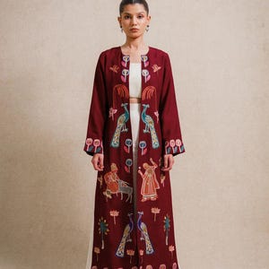 May include: A long, open-front burgundy duster coat featuring intricate multi-coloured embroidery of peacocks, floral motifs, and human figures. The garment is worn over a plain white crop top and wide-leg trousers, creating a layered ensemble.