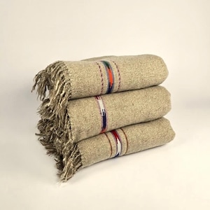 May include: A stack of three beige woven blankets with colorful striped accents. Each blanket has a decorative fringe on one end. The blankets are folded and stacked neatly, showcasing the intricate weave and design.