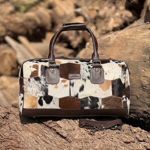 May include: A patchwork leather duffel bag with a brown leather handle and trim. The bag features a mix of brown, black, and white cowhide patches. Includes a detachable shoulder strap.