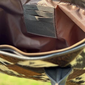 K&ouml;nnte beinhalten: Innenansicht einer Tragetasche aus Kuhfellleder. Die Tasche hat ein braun-wei&szlig;es Fleckenmuster auf der Au&szlig;enseite, mit dunkelbraunen Lederbes&auml;tzen und -griffen. Der Innenraum ist mit einem hellbraunen Stoff gef&uuml;ttert und enth&auml;lt einen dunkelbraunen Kartenhalter.