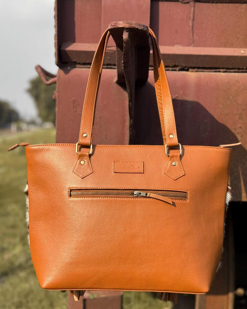 Pode incluir: Uma mala tote de couro castanho com um bolso frontal com z&iacute;per e duas al&ccedil;as superiores. A mala tem ferragens douradas e uma pequena etiqueta retangular. As laterais da mala t&ecirc;m um design branco e cinza.