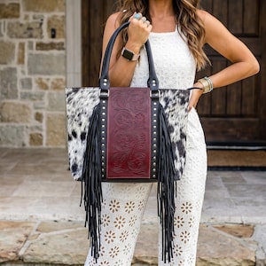 May include: A western-style tote bag with a cowhide pattern on the sides and a burgundy leather center panel. The bag features black fringe, silver studs, and black handles. The bag is a stylish accessory.